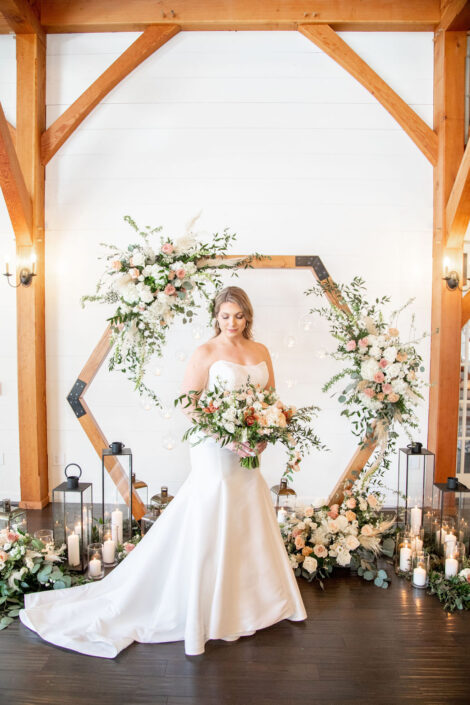 A bride standing in front of a wooden wedding arch with florals and candles.