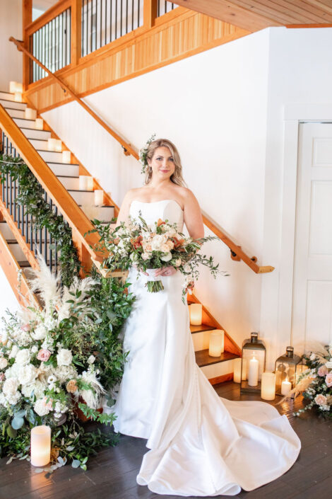 A bride standing in front of a staircase with a bouquet of greenery and candles.