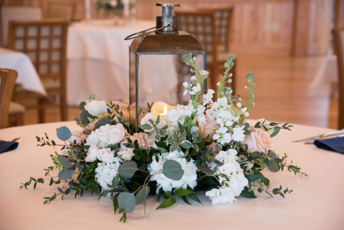 A lantern centerpiece with white flowers and greenery on a table.