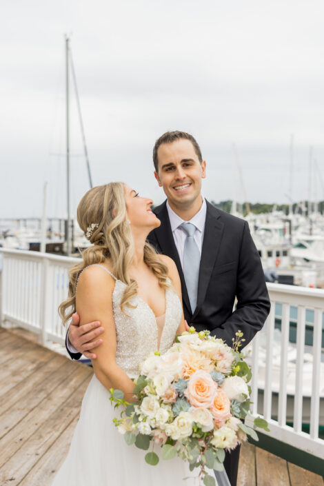 A bride and groom standing on a dock with boats in the background.