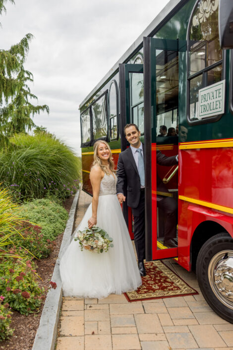 A bride and groom standing next to a red trolley bus.