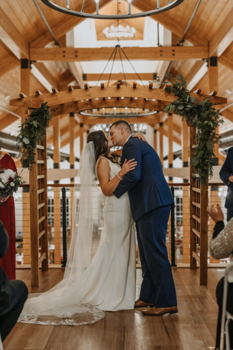 A bride and groom kiss during their wedding ceremony in a barn.